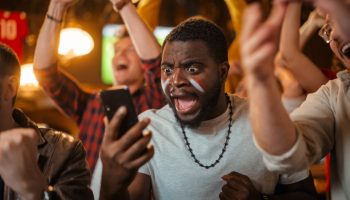 Portrait of an Excited Young Black Man Holding a Smartphone, Anxious About a Sports Bet