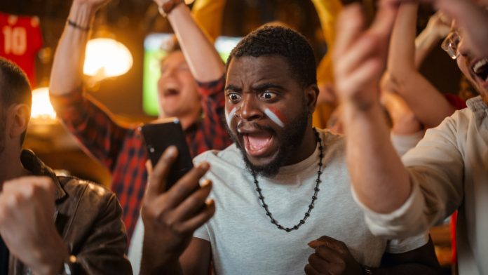 Portrait of an Excited Young Black Man Holding a Smartphone, Anxious About a Sports Bet